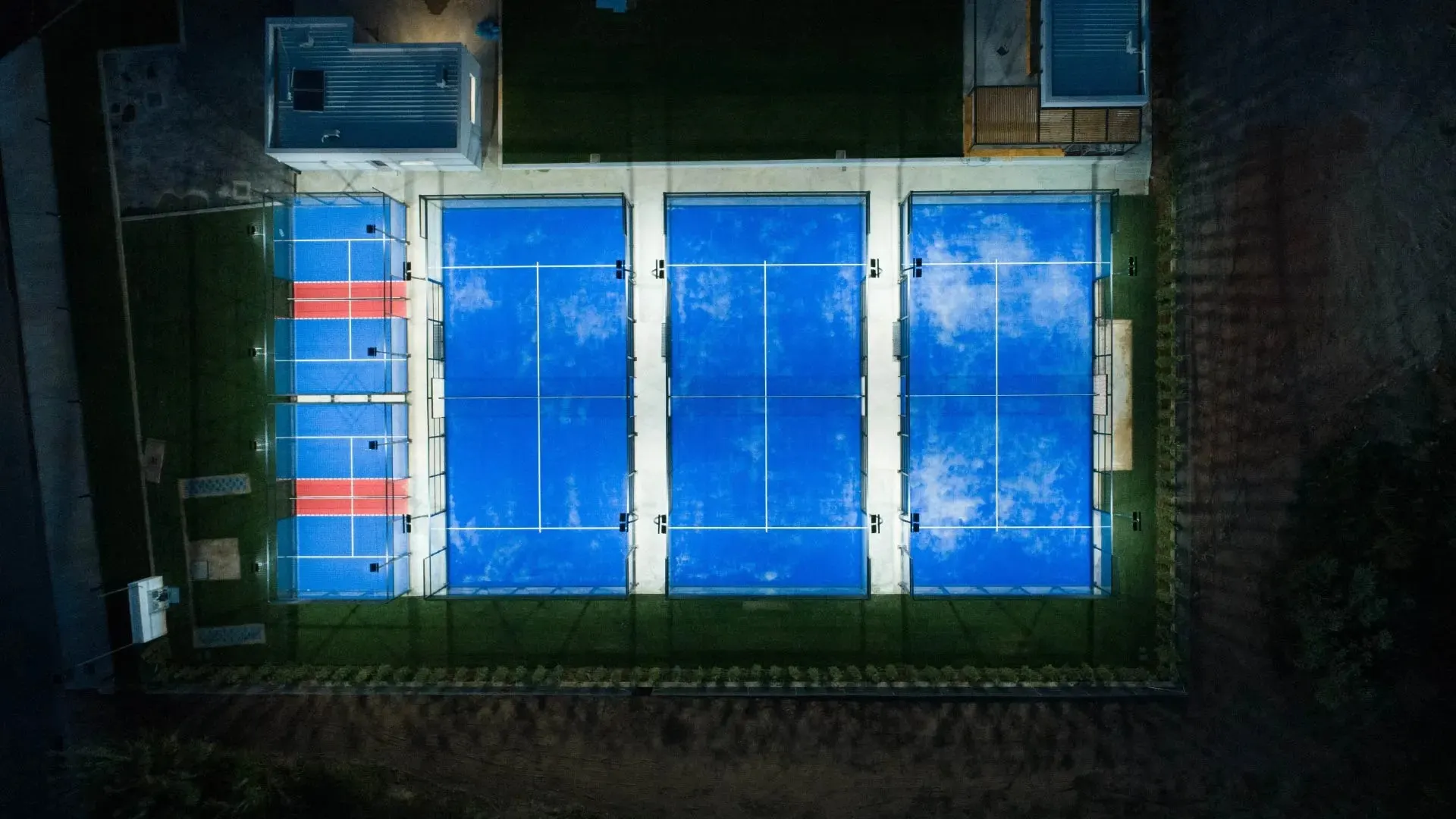 Aerial view of three floodlit padel courts and two smaller courts beside clubhouse buildings
