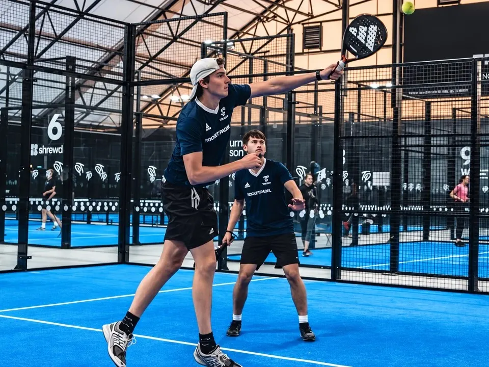 Two players in team kit play doubles on an indoor padel court